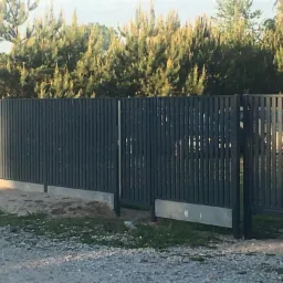 Nowoczesne, metalowe ogrodzenie panelowe w kolorze grafitowym z widoczną furtką i betonową podmurówką, na tle drzew iglastych.