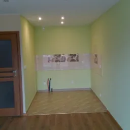 Wnętrze pokoju z zielonymi ścianami, częściowo położonymi panelami podłogowymi w dwóch odcieniach brązu, przygotowane do dalszych prac wykończeniowych, widoczne wystające rury instalacji.