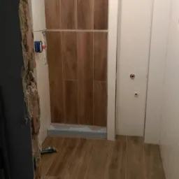 Łazienka w trakcie remontu: widoczne płytki imitujące drewno na podłodze i ścianie prysznica, częściowo wykończona ściana z białymi płytkami i otwór na drzwi, surowa ściana w lewym dolnym rogu...