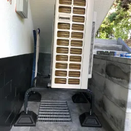 Zewnętrzna jednostka klimatyzacji zamontowana na metalowej podstawie z czarnymi stopkami, obok kratki ściekowej i schodów, z poziomnicą leżącą na schodach.