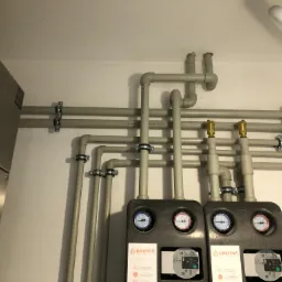 Szare rury instalacyjne z manometrami i pompami obiegowymi marki Warmit zamocowane do ściany, widok z dołu.