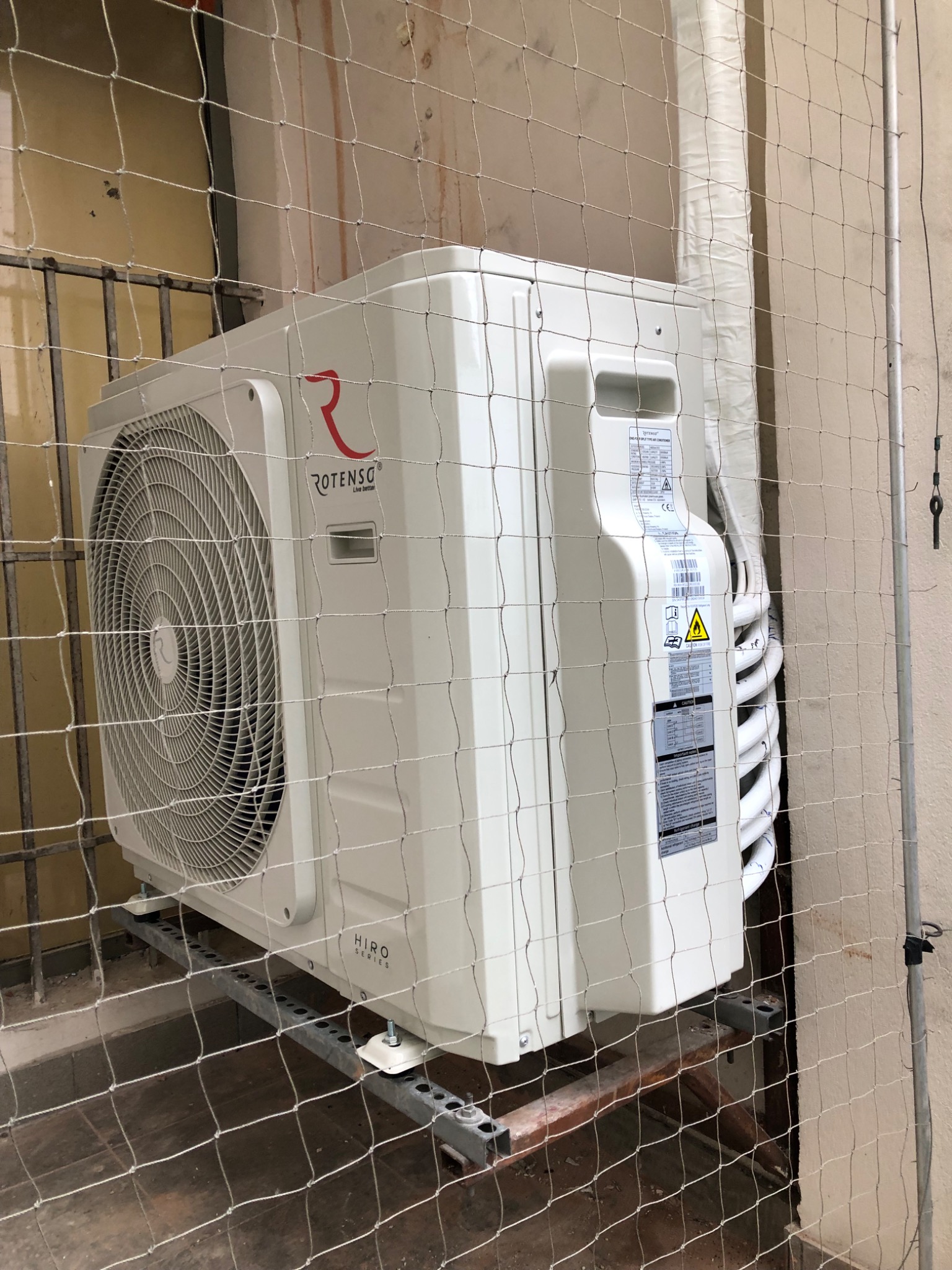 Zewnętrzna jednostka klimatyzacji Rotenso Hiro Series zamontowana na metalowej konstrukcji, widoczna przez siatkę zabezpieczającą.