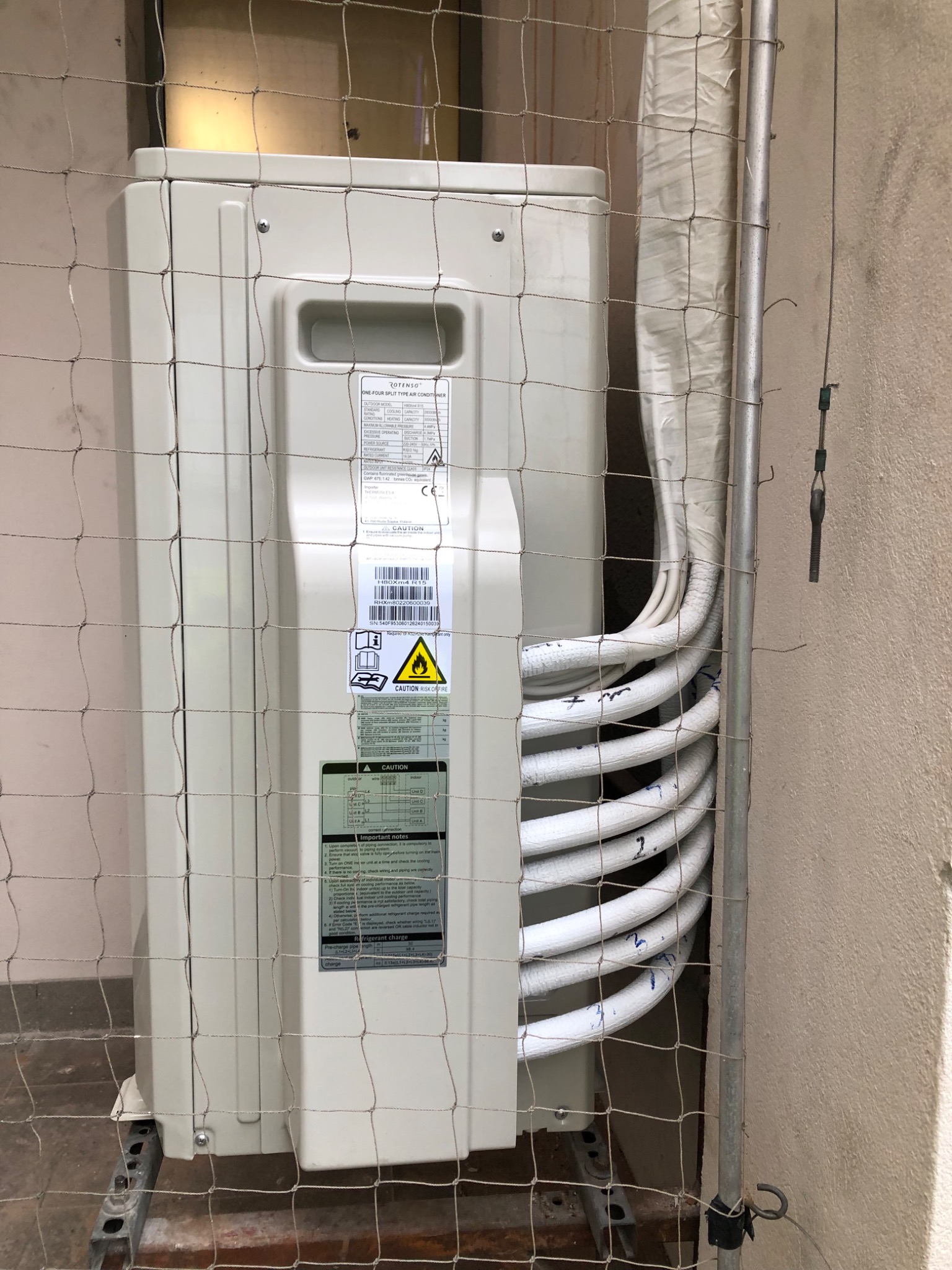 Zewnętrzna jednostka klimatyzacji Rotenso, widoczna przez siatkę ochronną, z podłączonymi rurami w izolacji, oznaczonymi numerami 1-4, zamontowana na metalowej konstrukcji przy ścianie budynku.