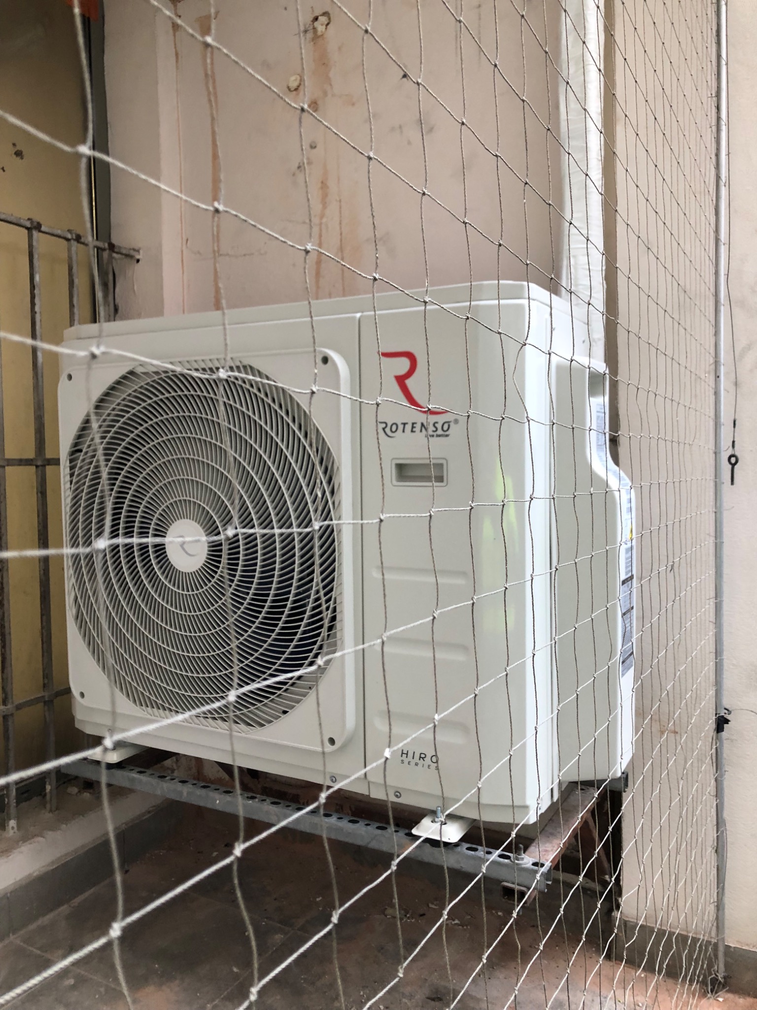 Zewnętrzna jednostka klimatyzacji Rotenso, zabezpieczona siatką ochronną, zamontowana na metalowej konstrukcji przy ścianie budynku.