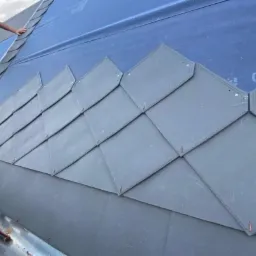 Układanie szarej dachówki łuskowej na membranie dachowej, widoczny fragment rynny i narzędzia