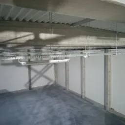 Wnętrze hali magazynowej z szarym betonowym podłożem, białymi ścianami z widocznymi słupami konstrukcyjnymi oraz zamontowanym systemem oświetlenia liniowego pod sufitem z blachy trapezowej.