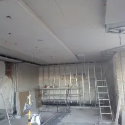 Wnętrze w trakcie remontu z widoczną konstrukcją metalową pod sufitem podwieszanym, częściowo zamontowane płyty gipsowo-kartonowe z widocznymi przewodami elektrycznymi i otworami na oświetlenie...