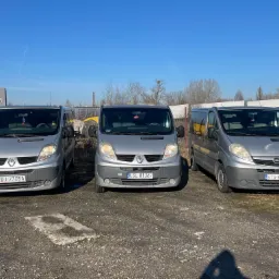 Trzy srebrne busy (Renault, Renault, Opel) zaparkowane obok siebie na żwirowym placu w słoneczny dzień. Każdy z nich ma inną tablicę rejestracyjną.
