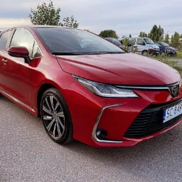 Czerwona Toyota Corolla hatchback z aluminiowymi felgami na tle parkingu z innymi pojazdami i zielenią.