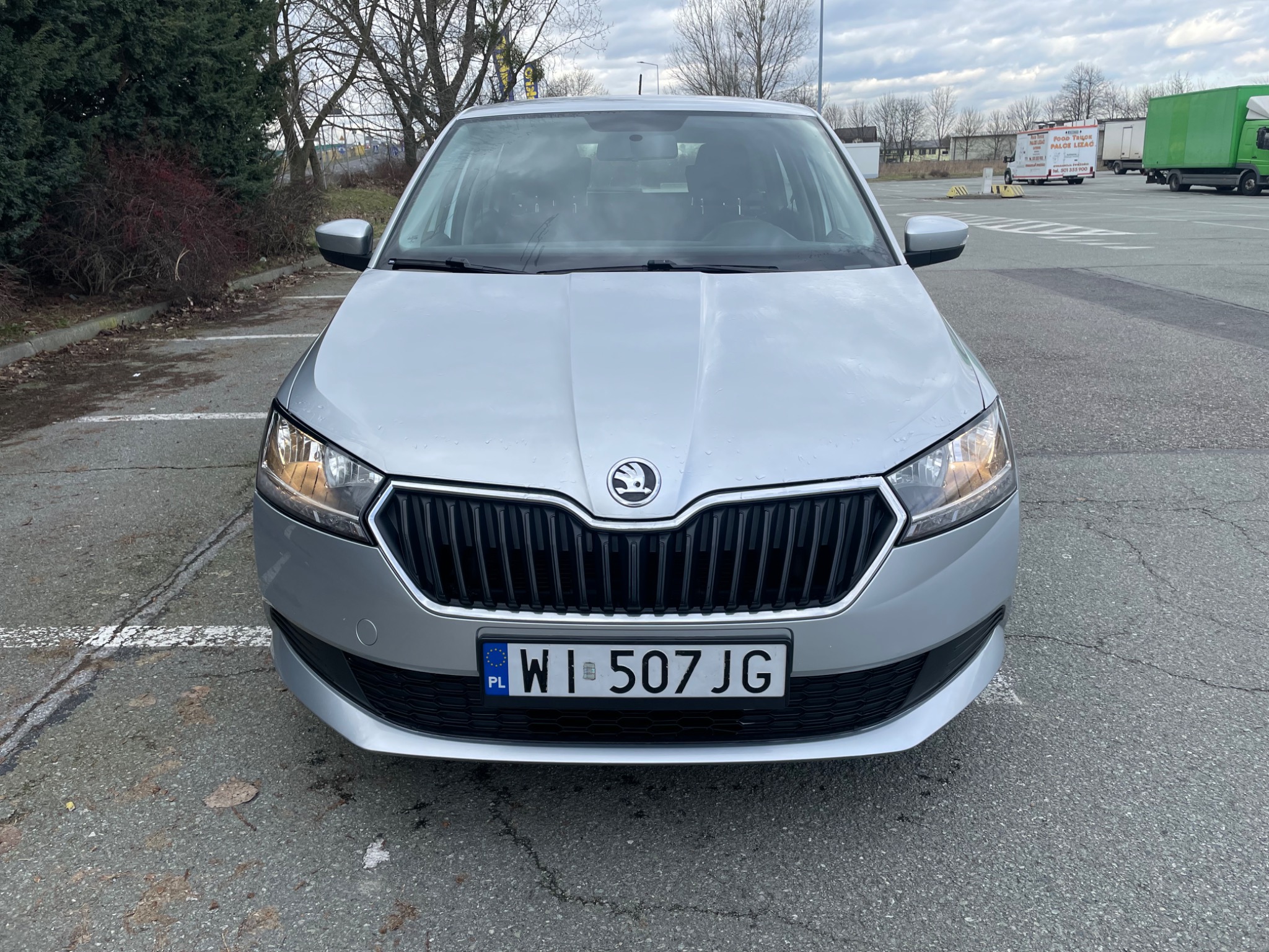 Srebrna Skoda Fabia, widok z przodu na parkingu, z widoczną tablicą rejestracyjną WI 507JG, na tle drzew i budynków.