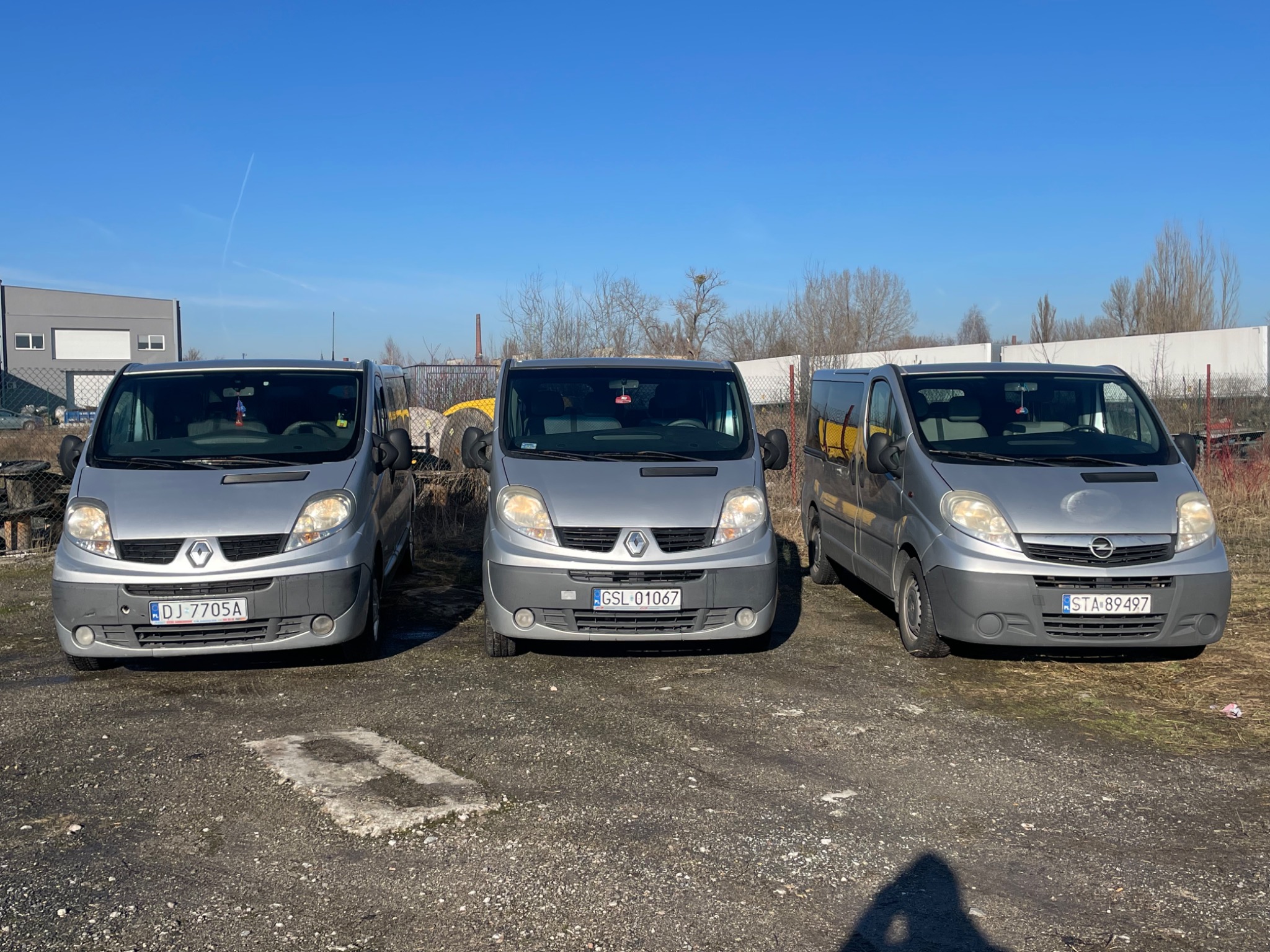 Trzy srebrne busy (Renault, Renault, Opel) zaparkowane obok siebie na żwirowym placu w słoneczny dzień. Każdy z nich ma inną tablicę rejestracyjną.