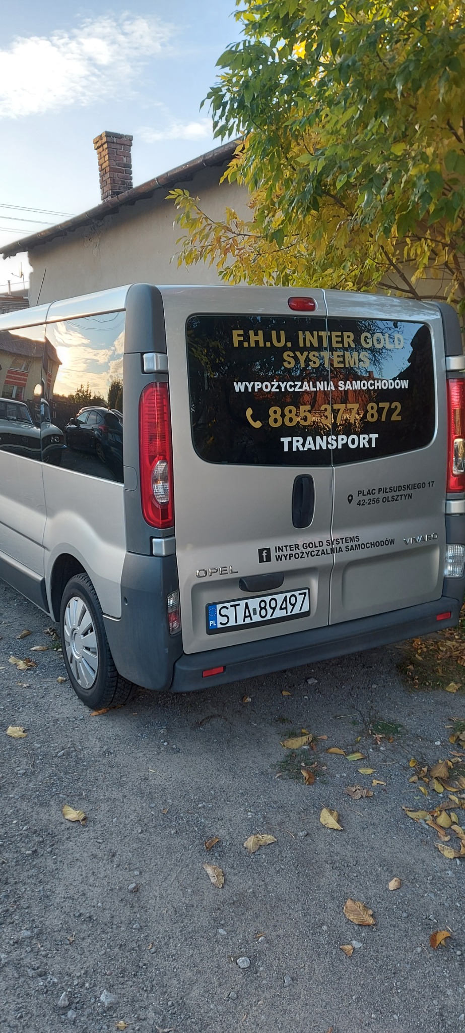 Szary Opel Vivaro z naklejkami reklamowymi firmy Inter Gold Systems, wypożyczalnia samochodów i transport, zaparkowany na żwirowym podłożu z opadłymi liśćmi, widok z tyłu, tablica rejestracyjna STA...