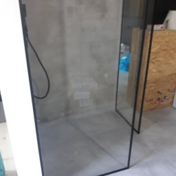 V-Glass - Nowoczesna kabina prysznicowa ze szklaną ścianą i czarnymi profilami, widoczna w trakcie montażu, z elementami instalacji w tle.