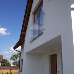 V-Glass - Nowoczesny balkon z przezroczystą taflą szkła mocowaną punktowo do białej ściany budynku, widoczne przewody elektryczne wystające ze ściany wewnątrz.