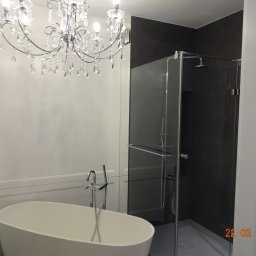 V-Glass - Nowoczesna łazienka z owalną wanną wolnostojącą, szklaną kabiną prysznicową i ozdobnym żyrandolem kryształowym, oświetlona ciepłym światłem.