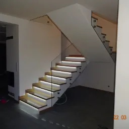 Nowoczesne schody drewniane z podświetleniem LED i szklaną balustradą, widoczne mocowania szkła, minimalistyczny design wnętrza.