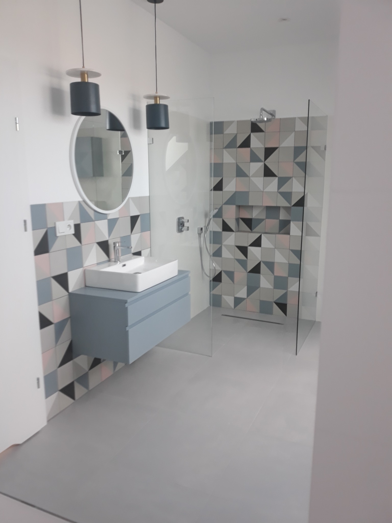 Nowoczesna łazienka z kabiną prysznicową ze szkła i umywalką na szafce w kolorze gołębim, wykończona geometrycznymi płytkami w odcieniach szarości, bieli i różu.