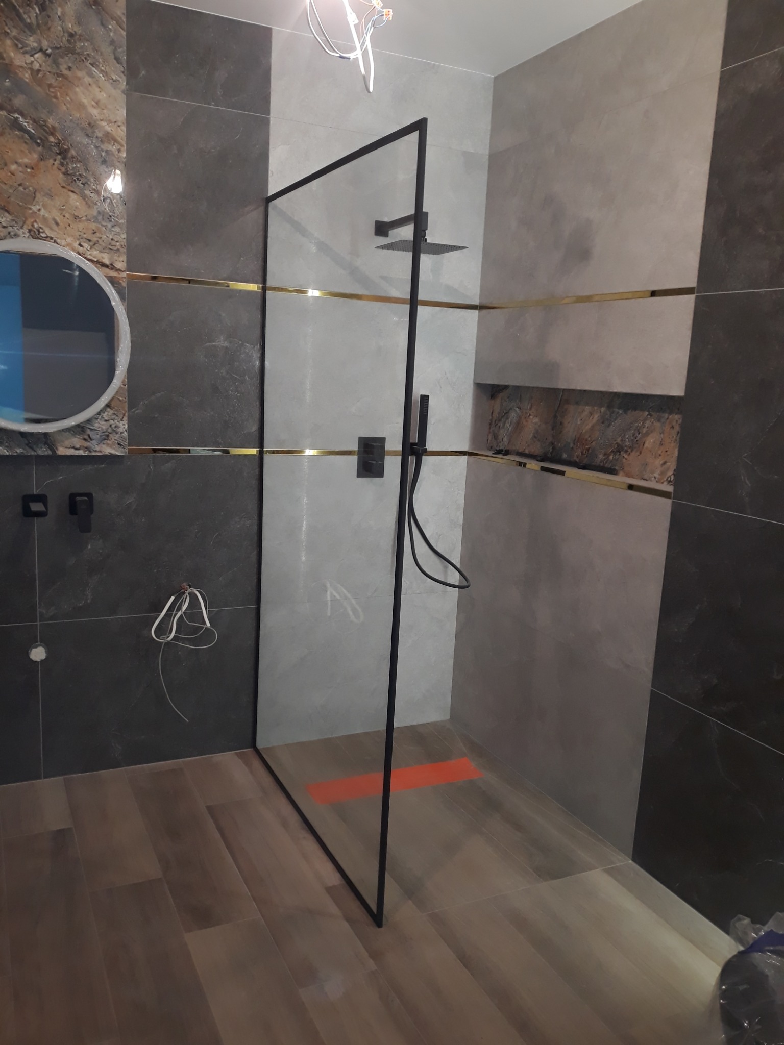 Nowoczesna kabina prysznicowa typu walk-in z czarnymi profilami i deszczownicą, wykończona szarymi płytkami z dekoracyjnym złotym paskiem, widoczne niedokończone podłączenia elektryczne.