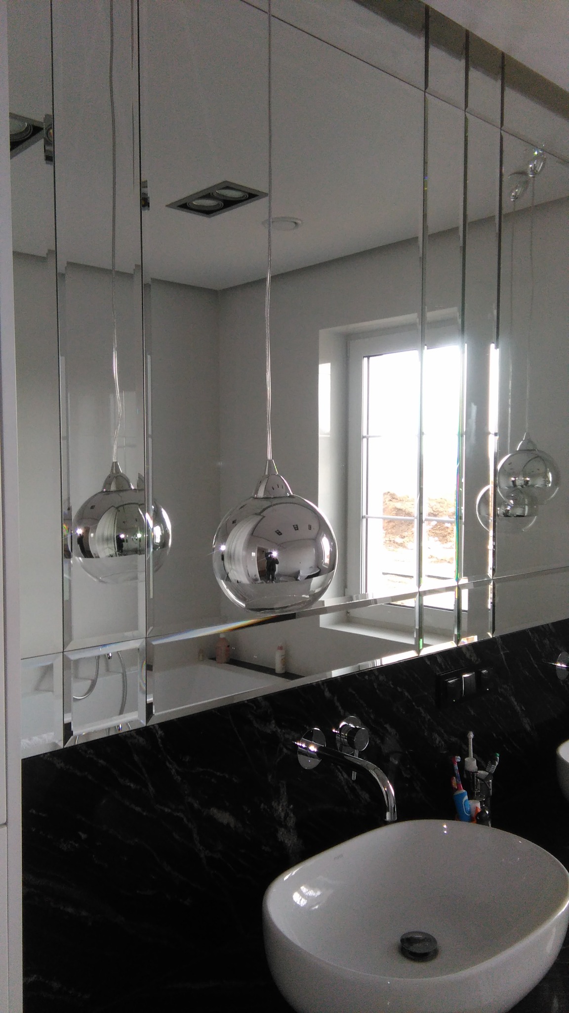 Łazienka z lustrem z pionowych szklanych pasów z fazowanymi krawędziami, odbijającym wiszące chromowane lampy kuliste, umywalkę nablatową i okno z widokiem na plac budowy.