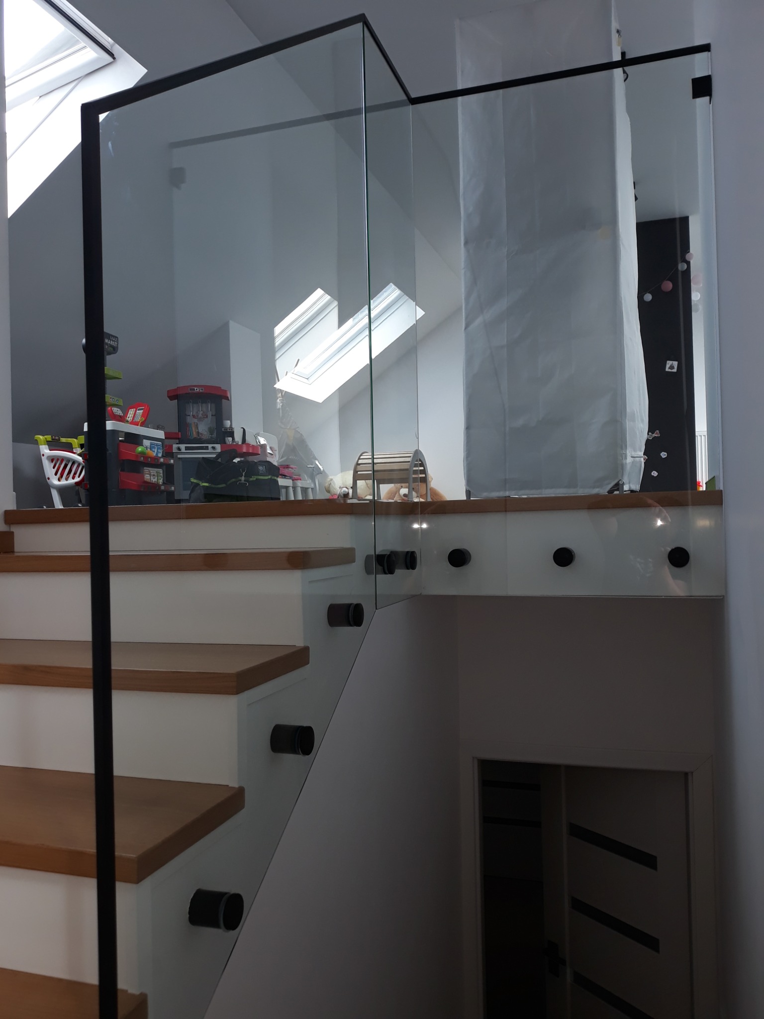 Szklana barierka z czarnymi elementami mocującymi, zabezpieczająca schody na poddaszu w Sokołowie Podlaskim, widok na zabawki dziecięce za szybą.