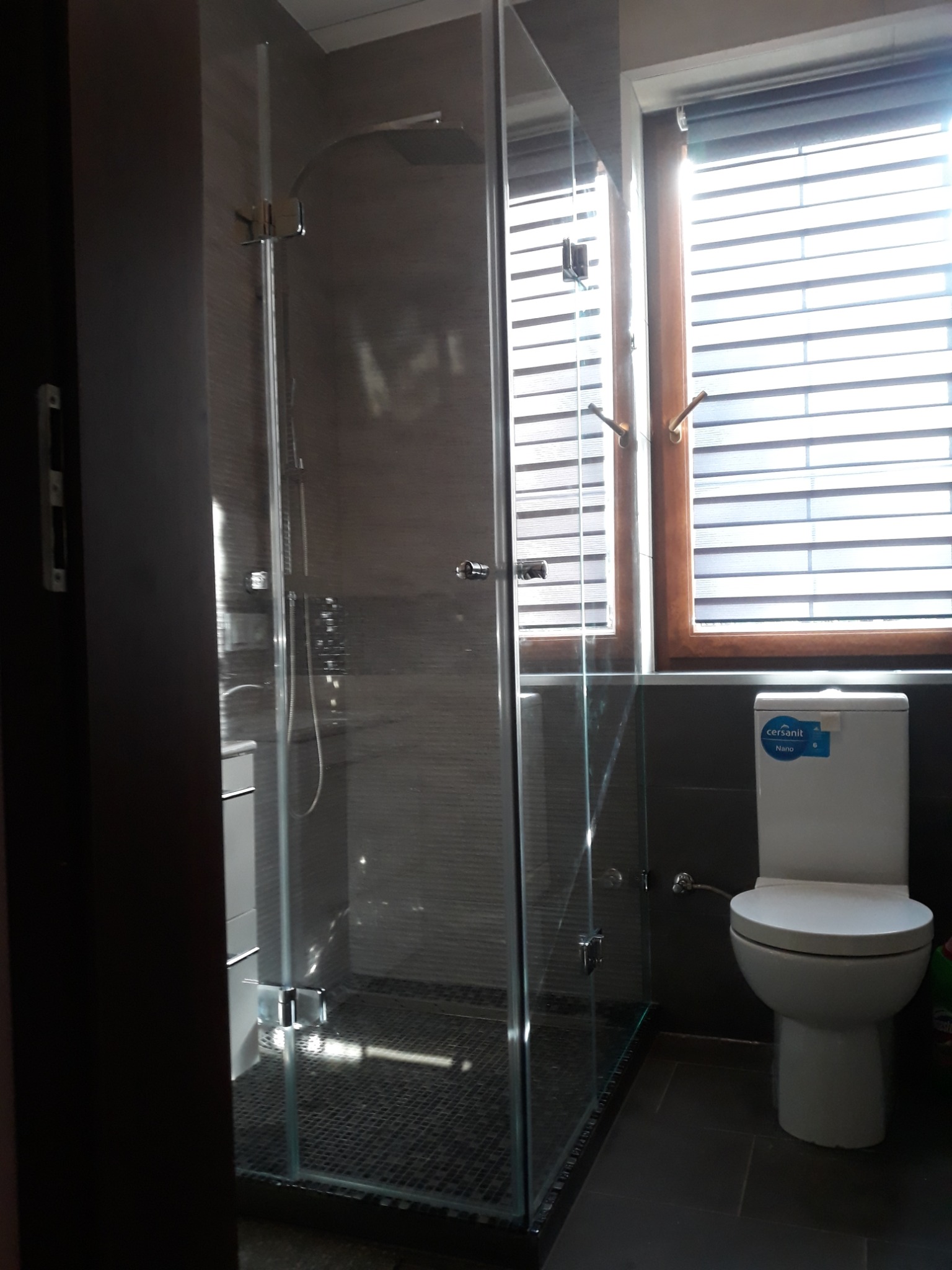 Szklana kabina prysznicowa z chromowanymi zawiasami i deszczownicą, widoczna w łazience z szarymi płytkami i oknem z żaluzjami.