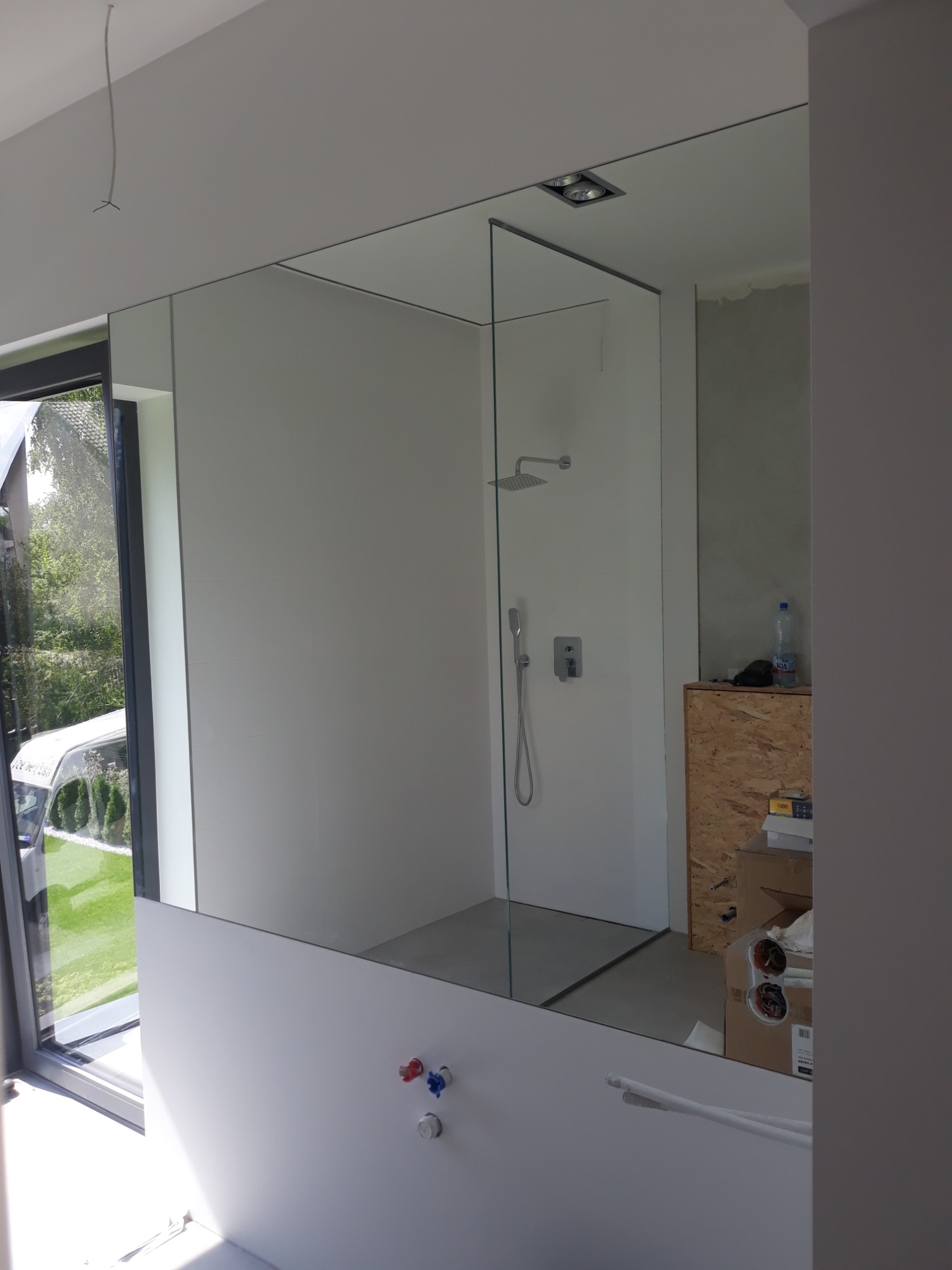 Nowoczesna łazienka z kabiną prysznicową oddzieloną szklaną ścianką, widoczne zawory i lustro na ścianie, w tle okno z widokiem na zieleń i częściowo zakryta kartonami szafka.