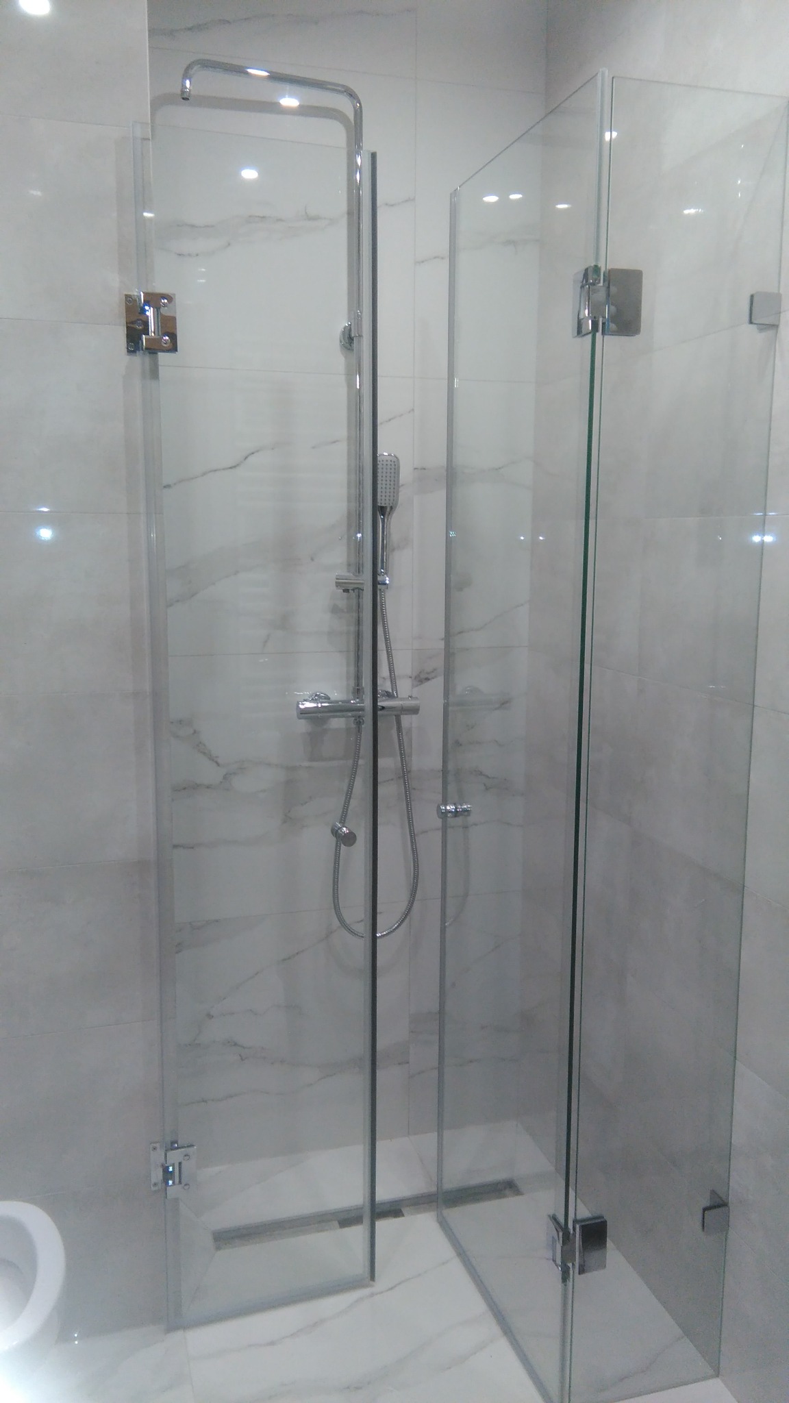 Szklana kabina prysznicowa z chromowanymi zawiasami i odpływem liniowym w posadzce, na tle marmurowych płytek.