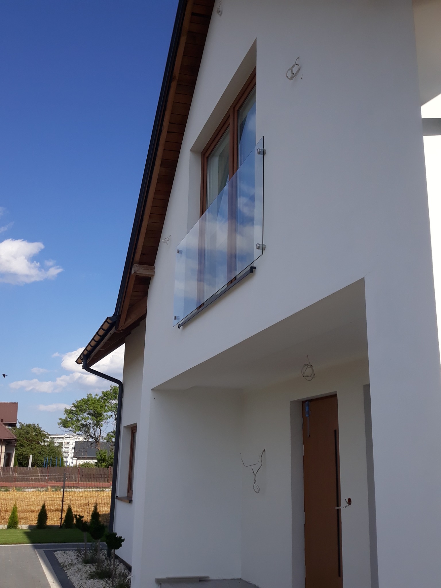 Nowoczesny balkon z przezroczystą taflą szkła mocowaną punktowo do białej ściany budynku, widoczne przewody elektryczne wystające ze ściany wewnątrz.