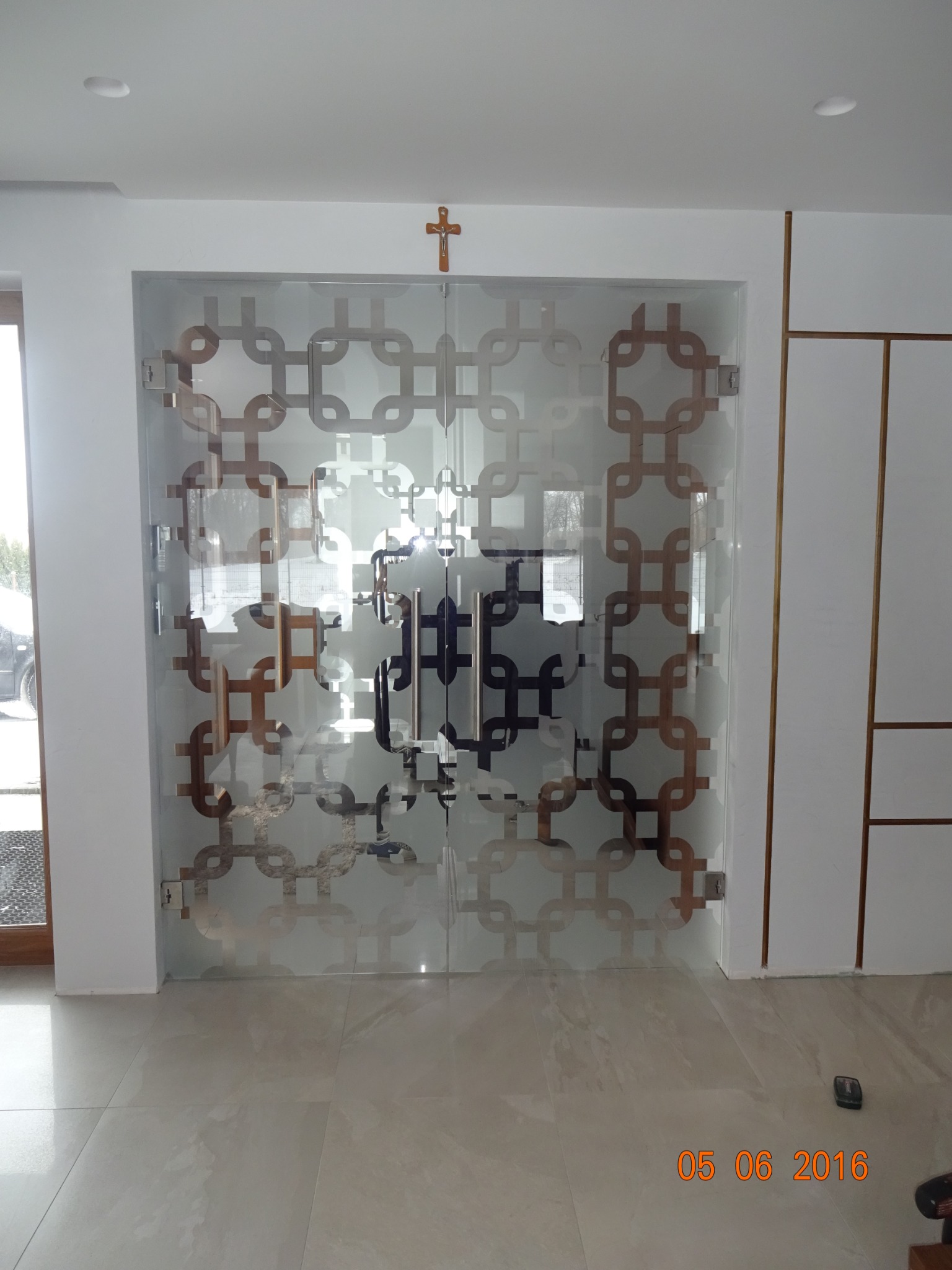 Szklane drzwi z geometrycznym wzorem w kościele, widoczny krzyż nad nimi, jasne płytki na podłodze.
