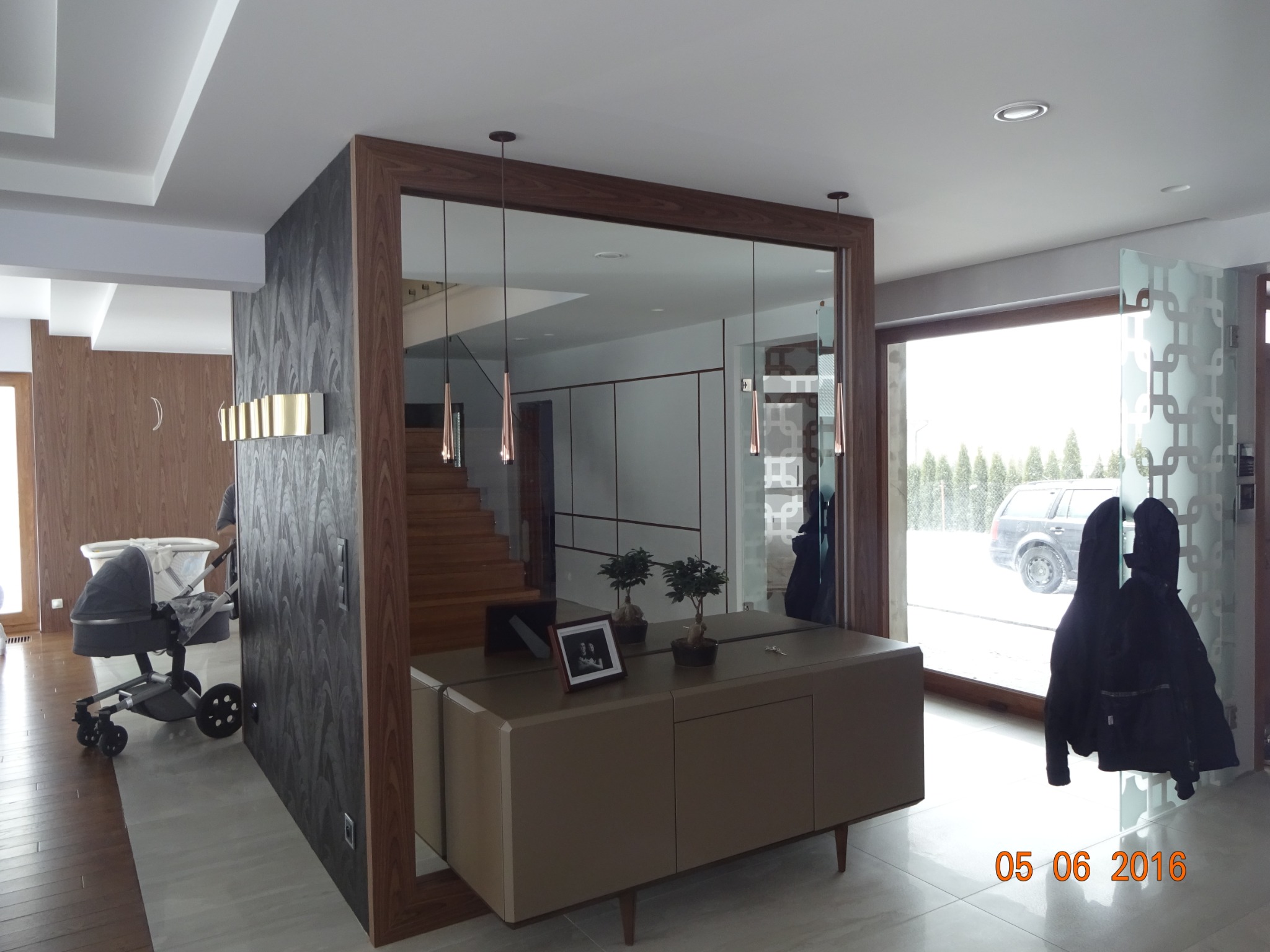 Nowoczesne wnętrze domu z lustrzaną ścianą w drewnianej ramie, wiszącymi lampami i komodą, widoczne schody i wózek dziecięcy.