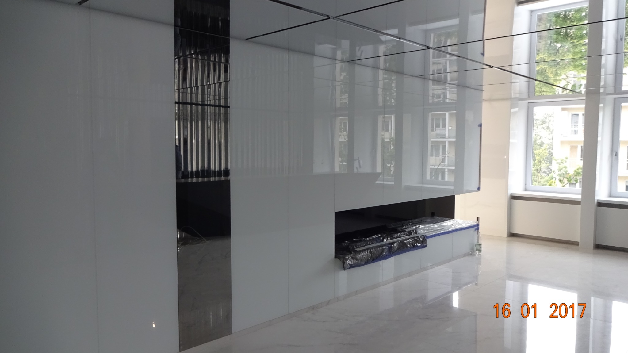 Białe, lakierowane panele ścienne z wbudowaną wnęką i lustrzanym panelem, odbijające światło z okien w przestronnym wnętrzu.