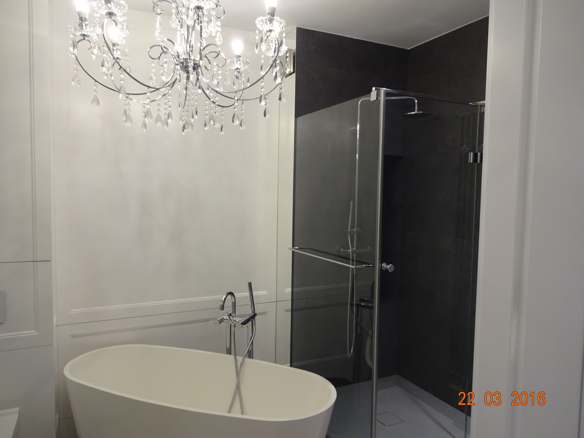 Nowoczesna łazienka z owalną wanną wolnostojącą, szklaną kabiną prysznicową i ozdobnym żyrandolem kryształowym, oświetlona ciepłym światłem.