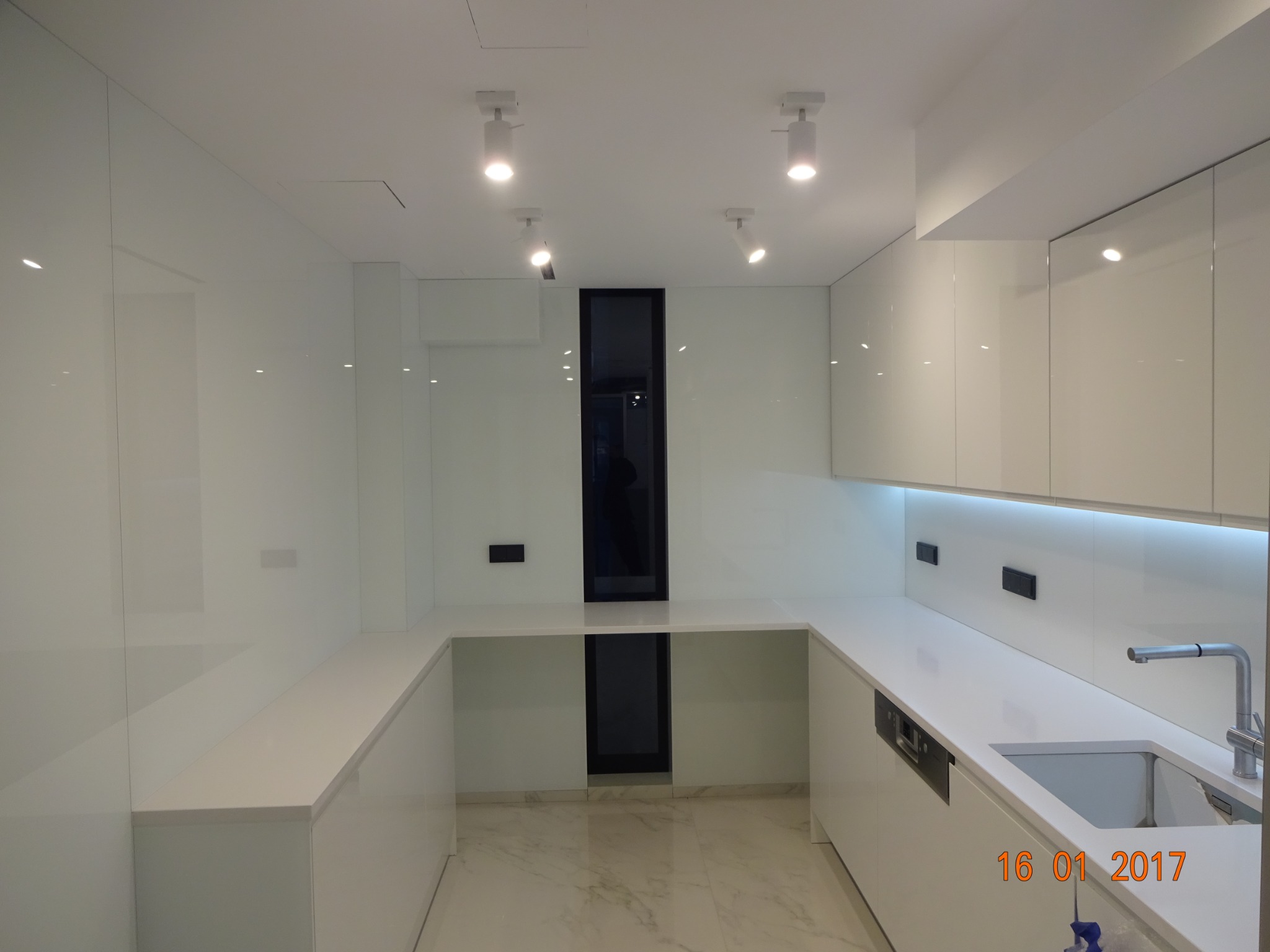 Nowoczesna, minimalistyczna kuchnia z białymi, błyszczącymi frontami szafek i blatami, oświetlona punktowymi lampami sufitowymi i ledowym podświetleniem podszafkowym, z marmurową posadzką i czarnym...