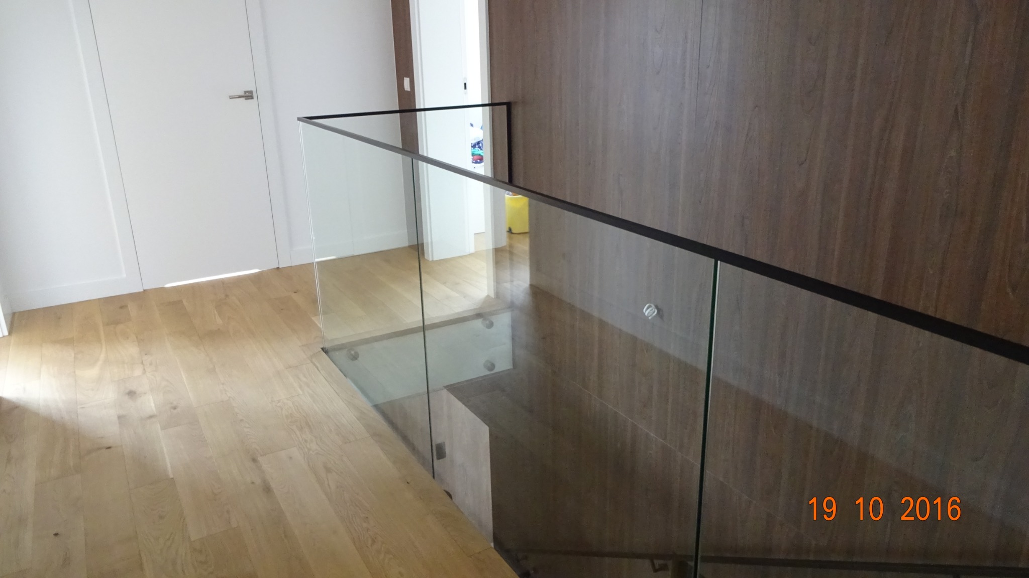 Szklana bariera schodowa z czarną ramą, widok z góry na drewniane schody i podłogę, jasne wnętrze z białymi drzwiami.