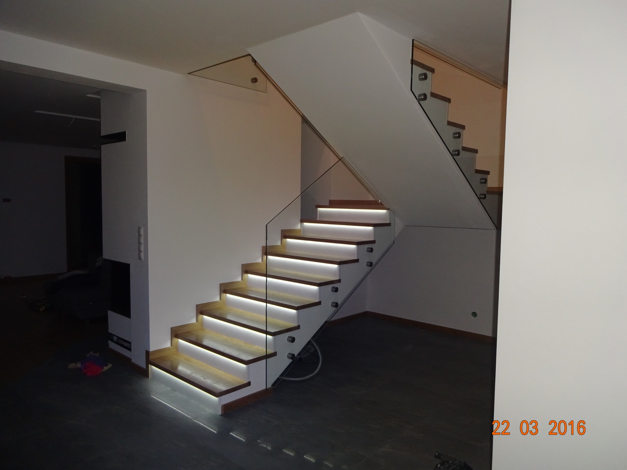 Nowoczesne schody drewniane z podświetleniem LED i szklaną balustradą, widoczne mocowania szkła, minimalistyczny design wnętrza.