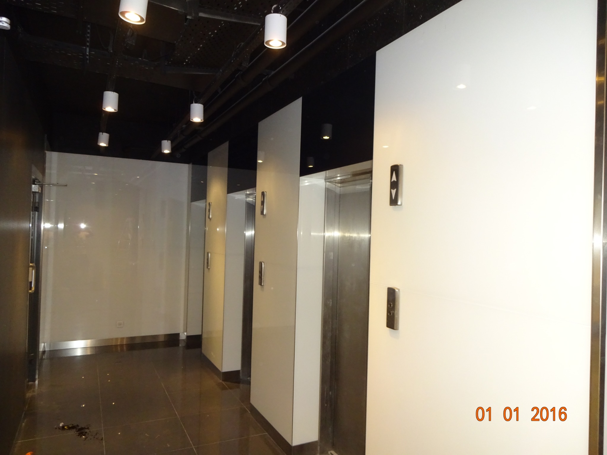 Wnętrze budynku z trzema windami z białymi panelami i metalowymi drzwiami, widoczne oświetlenie sufitowe i ciemna podłoga.