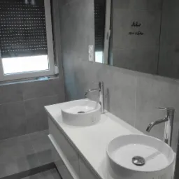 Nowoczesna łazienka z dwoma okrągłymi umywalkami nablatowymi, chromowanymi bateriami, dużym lustrem z odręcznym napisem i szarymi płytkami na ścianach i podłodze.