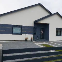 Nowoczesny dom jednorodzinny z szarą elewacją, ciemną stolarką okienną i brukowanym podjazdem z elementami trawnika, widziany zza metalowego ogrodzenia.