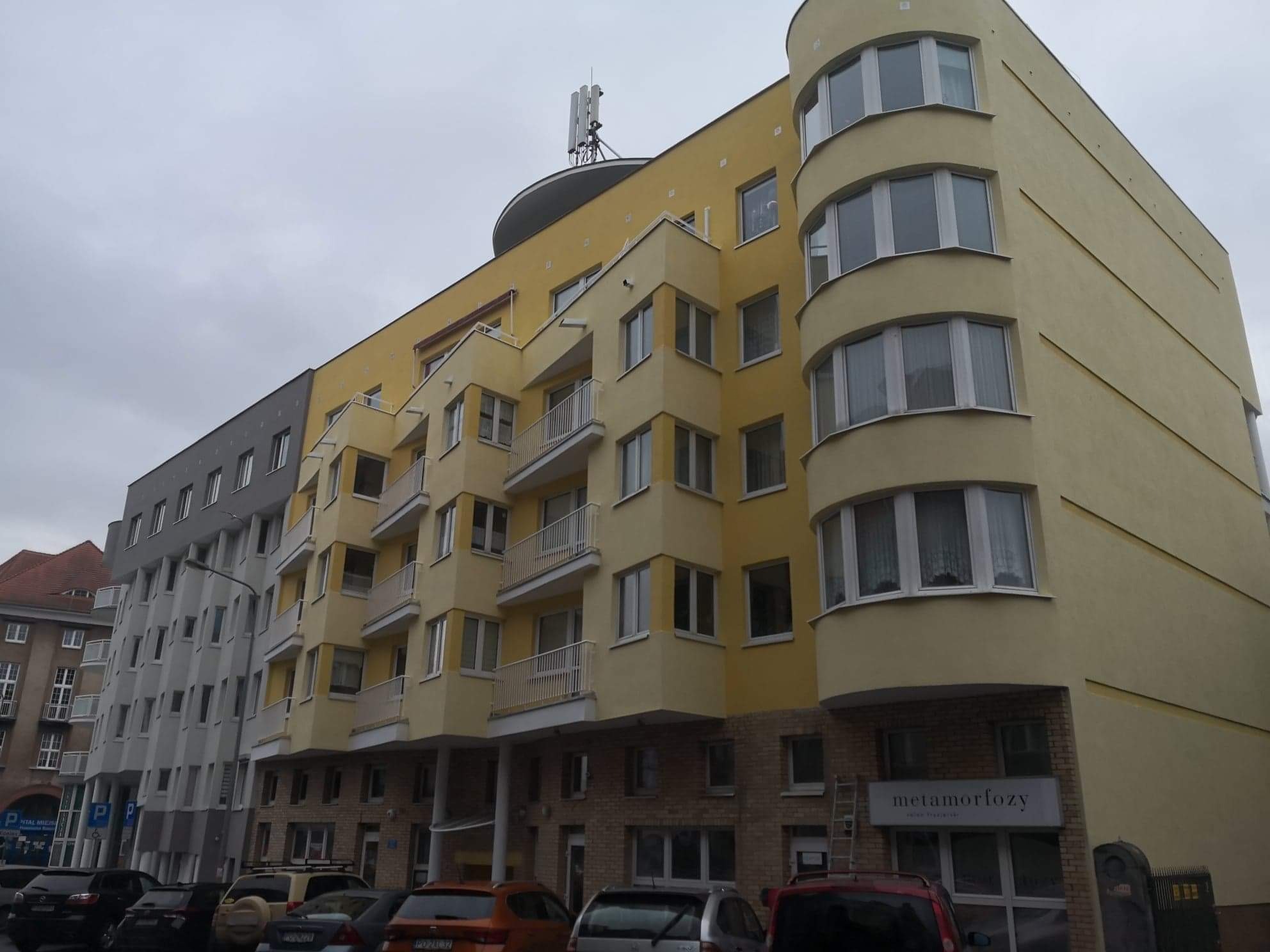 Odnowiony budynek mieszkalny z żółtą elewacją i balkonami, widoczna antena na dachu i zaparkowane samochody na pierwszym planie.