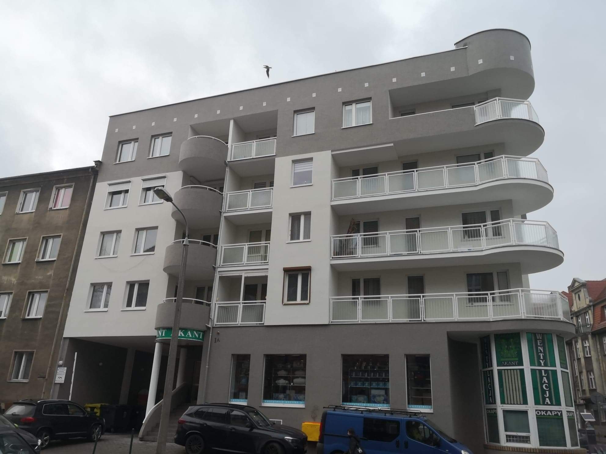 Szaro-biały budynek mieszkalny z zaokrąglonymi balkonami i widocznym szyldem 'Akant Wenty-Lacja Okapy' na parterze, z zaparkowanymi samochodami i przelatującym ptakiem na tle pochmurnego nieba.