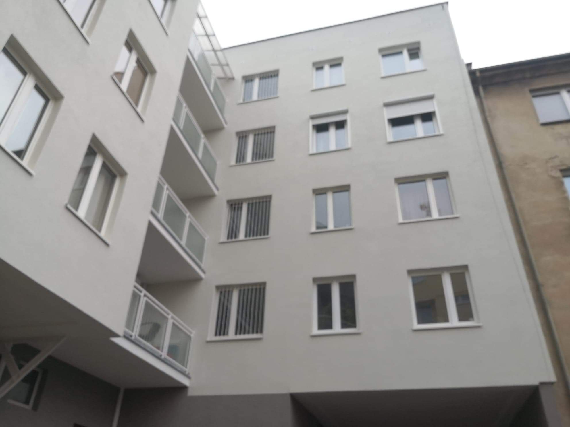 Elewacja odnowionego budynku mieszkalnego z balkonami i kratami w oknach, widok z dołu na szaro-białą fasadę.