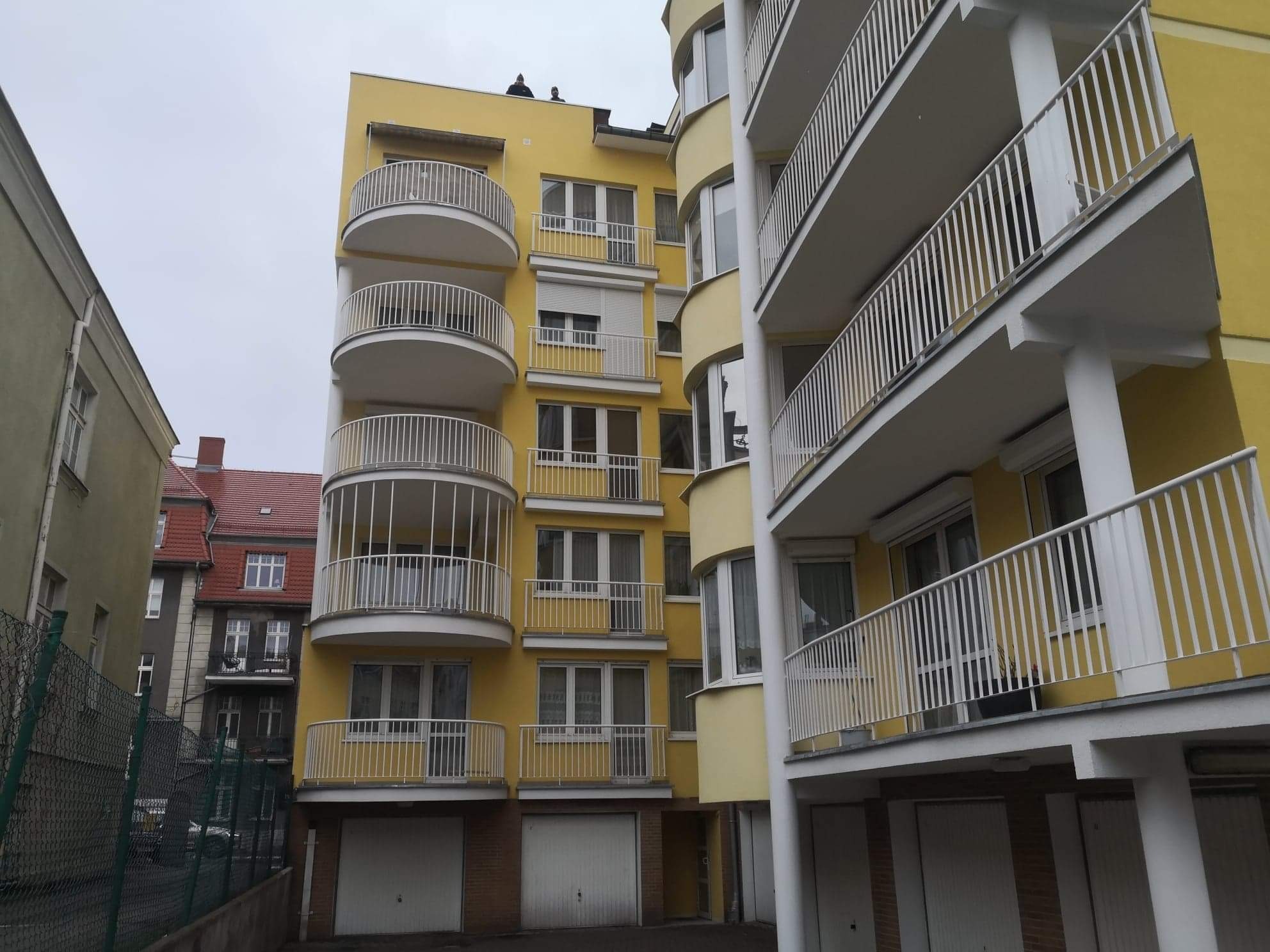 Żółty budynek mieszkalny z zaokrąglonymi balkonami i białymi balustradami, widok z dołu na pochmurne niebo. Na dachu budynku widoczne dwie osoby.