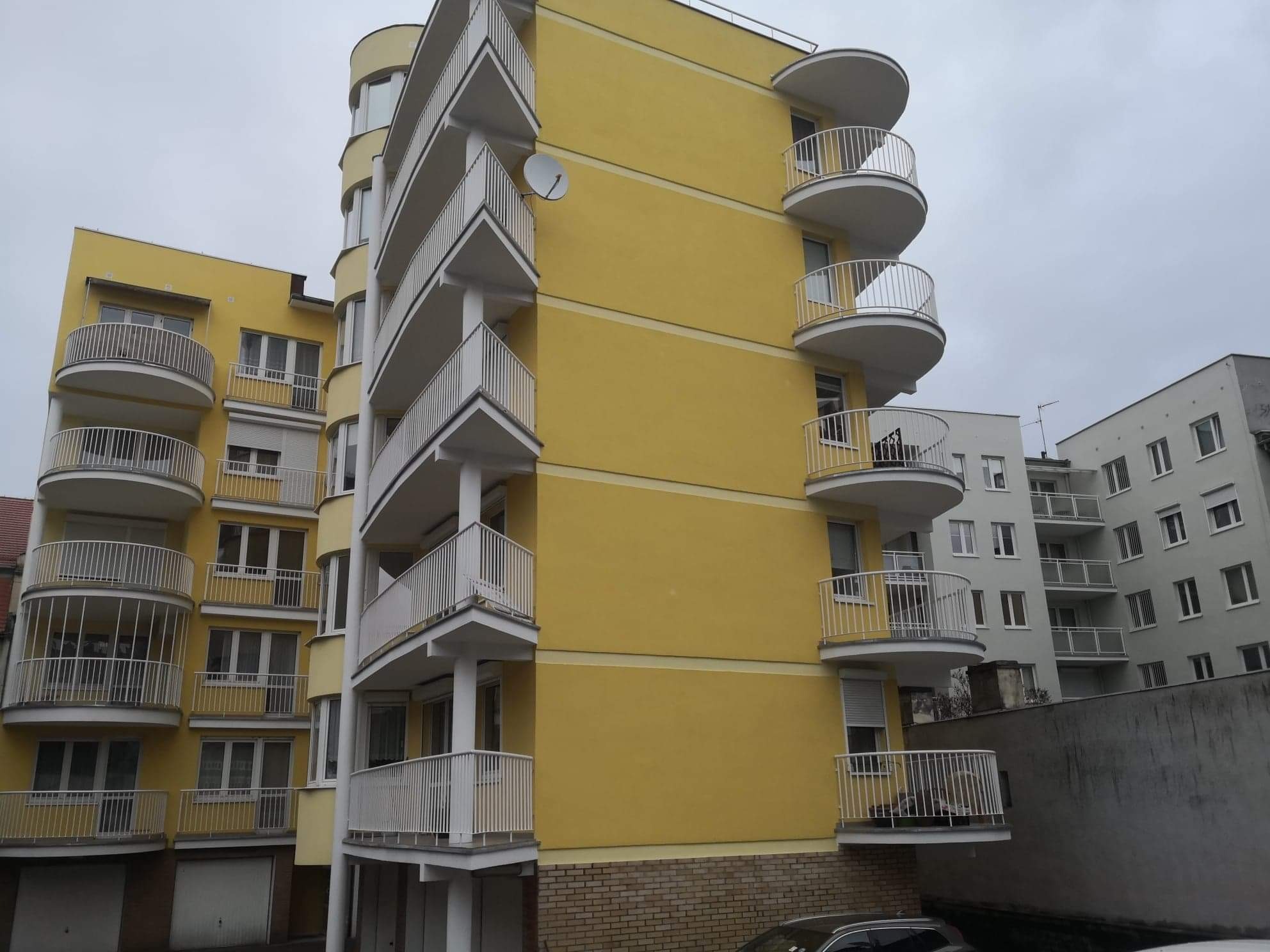 Żółty budynek mieszkalny z białymi, okrągłymi balkonami i anteną satelitarną na elewacji, widok z dołu w pochmurny dzień.