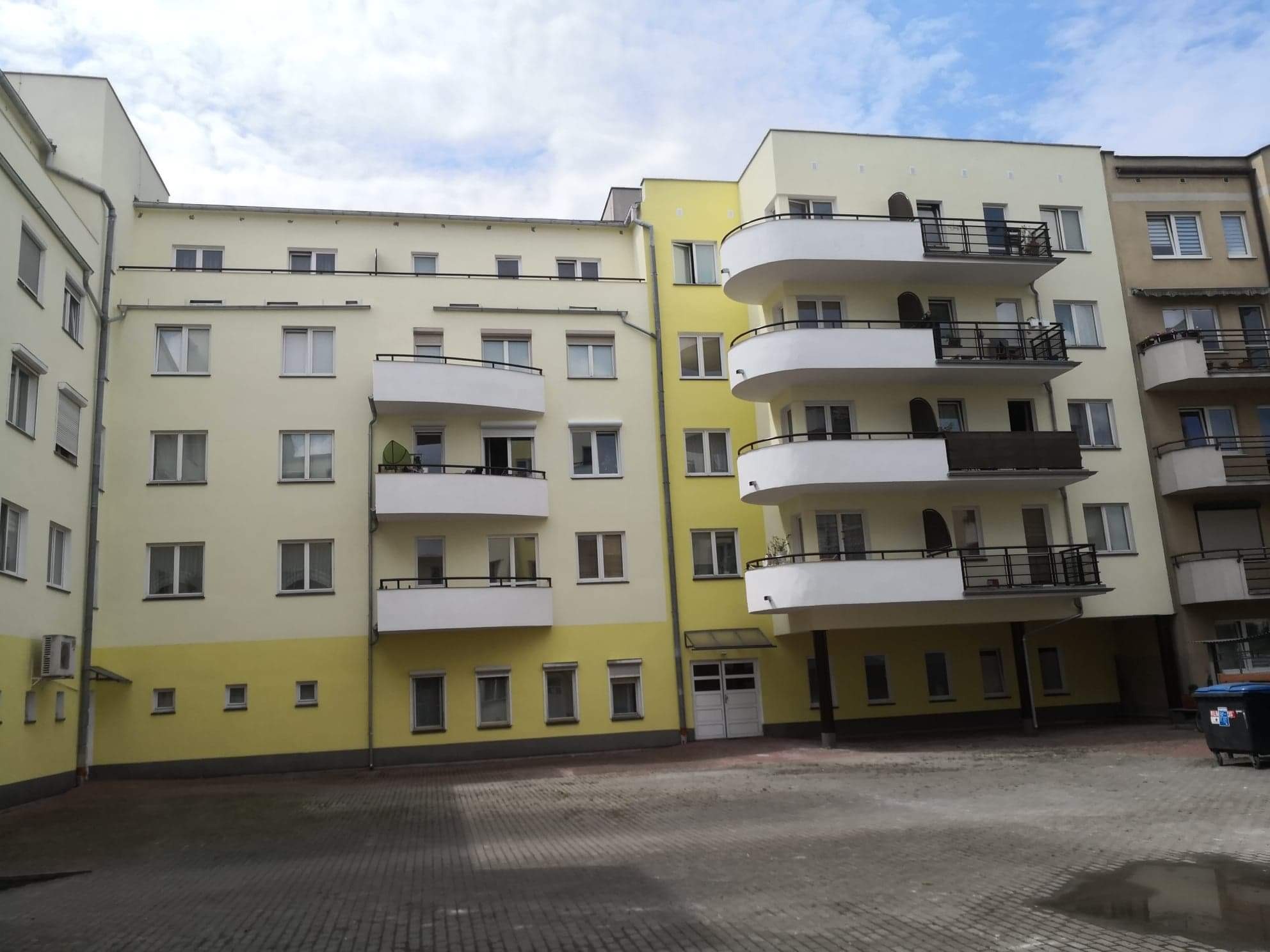 Fasada odnowionego budynku wielorodzinnego z balkonami w odcieniach żółci i bieli, widoczna z poziomu dziedzińca wyłożonego kostką.