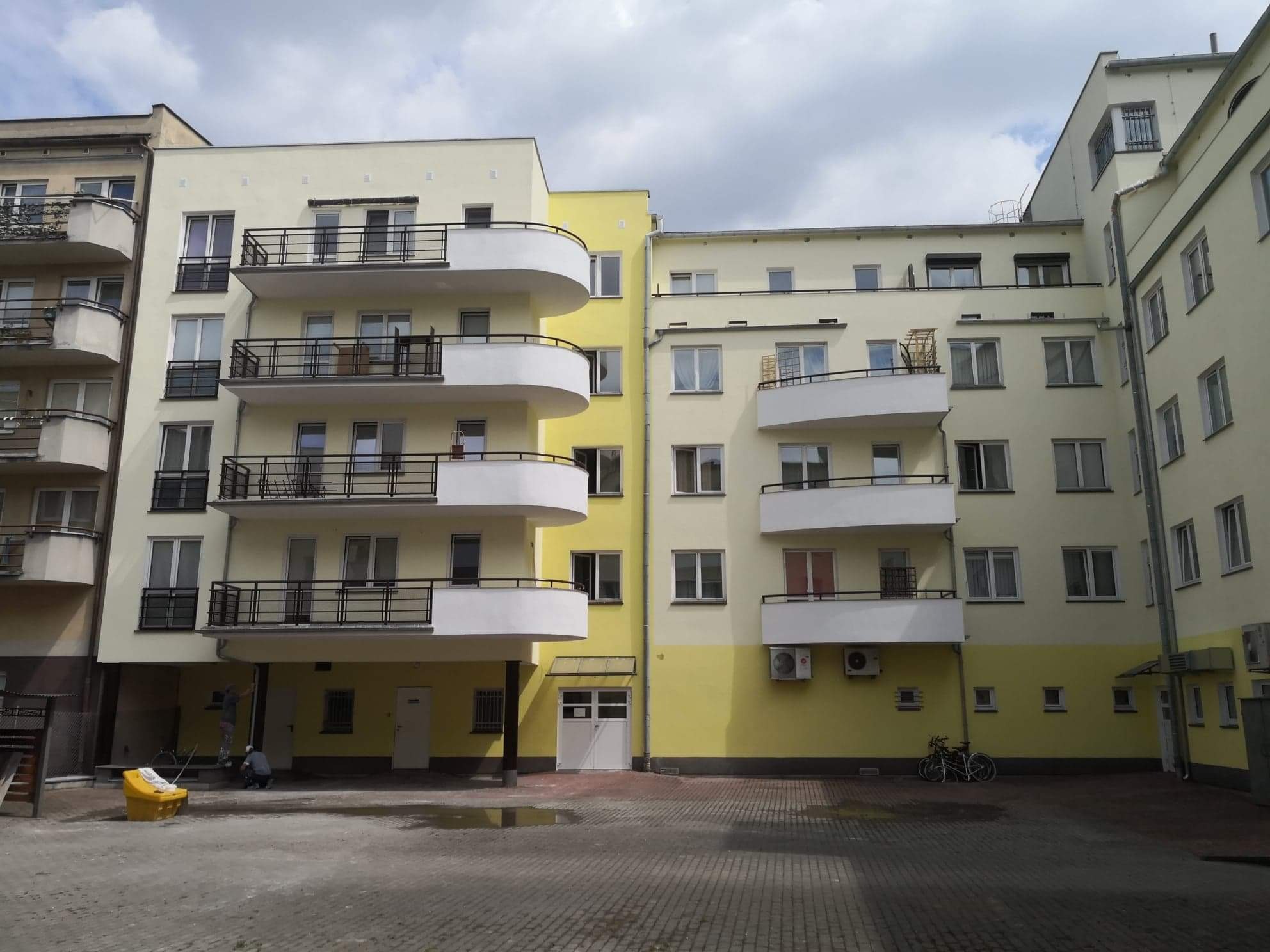 Prace malarskie na elewacji budynku wielorodzinnego w trakcie realizacji, widoczni dwaj pracownicy, żółta farba, balkony z czarnymi barierkami, kostka brukowa na dziedzińcu