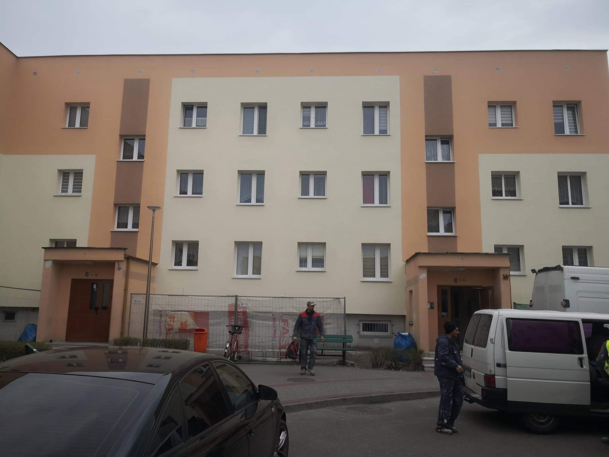 Pracownicy budowlani przed odnowionym, wielobarwnym budynkiem mieszkalnym z białymi oknami i ciemnym dachem samochodu na pierwszym planie.