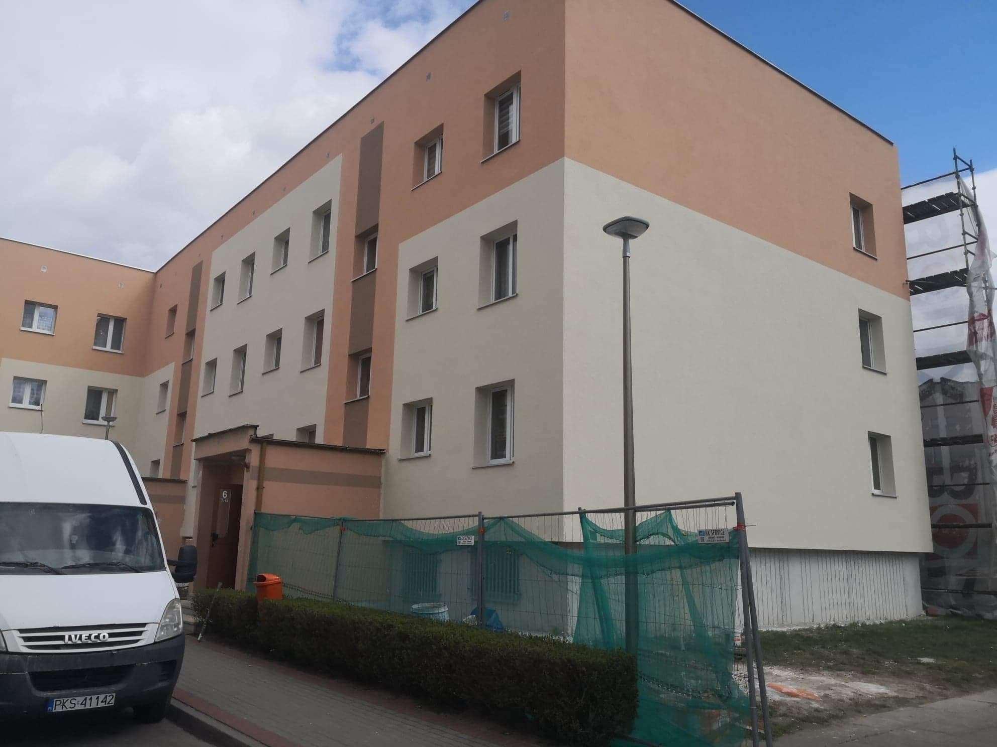 Odnowiony budynek wielorodzinny z elewacją w odcieniach beżu i kremu, zabezpieczony siatką ochronną i barierkami, z zaparkowanym busem z przodu.