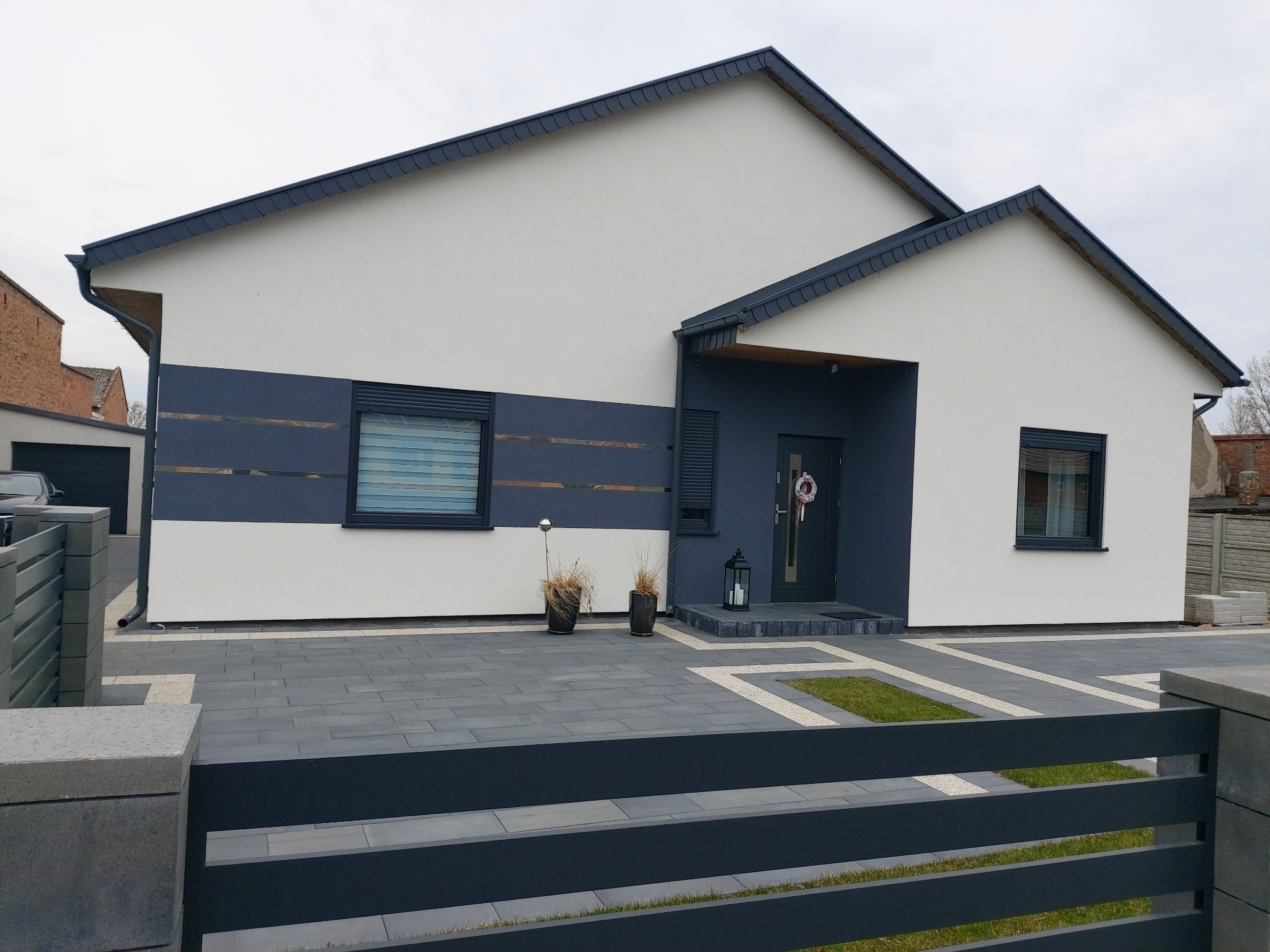 Nowoczesny dom jednorodzinny z szarą elewacją, ciemną stolarką okienną i brukowanym podjazdem z elementami trawnika, widziany zza metalowego ogrodzenia.