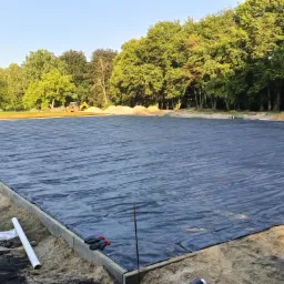 Przygotowany teren pod budowę z rozłożoną czarną geowłókniną, widoczne obrzeża betonowe, rura PCV, oraz koparka w oddali na tle drzew