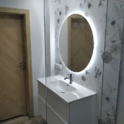 Nowoczesna łazienka z umywalką nablatową na szafce z szufladami, okrągłym lustrem z podświetleniem LED i płytkami z motywem kwiatowym na ścianie, kabina prysznicowa ze szkła.